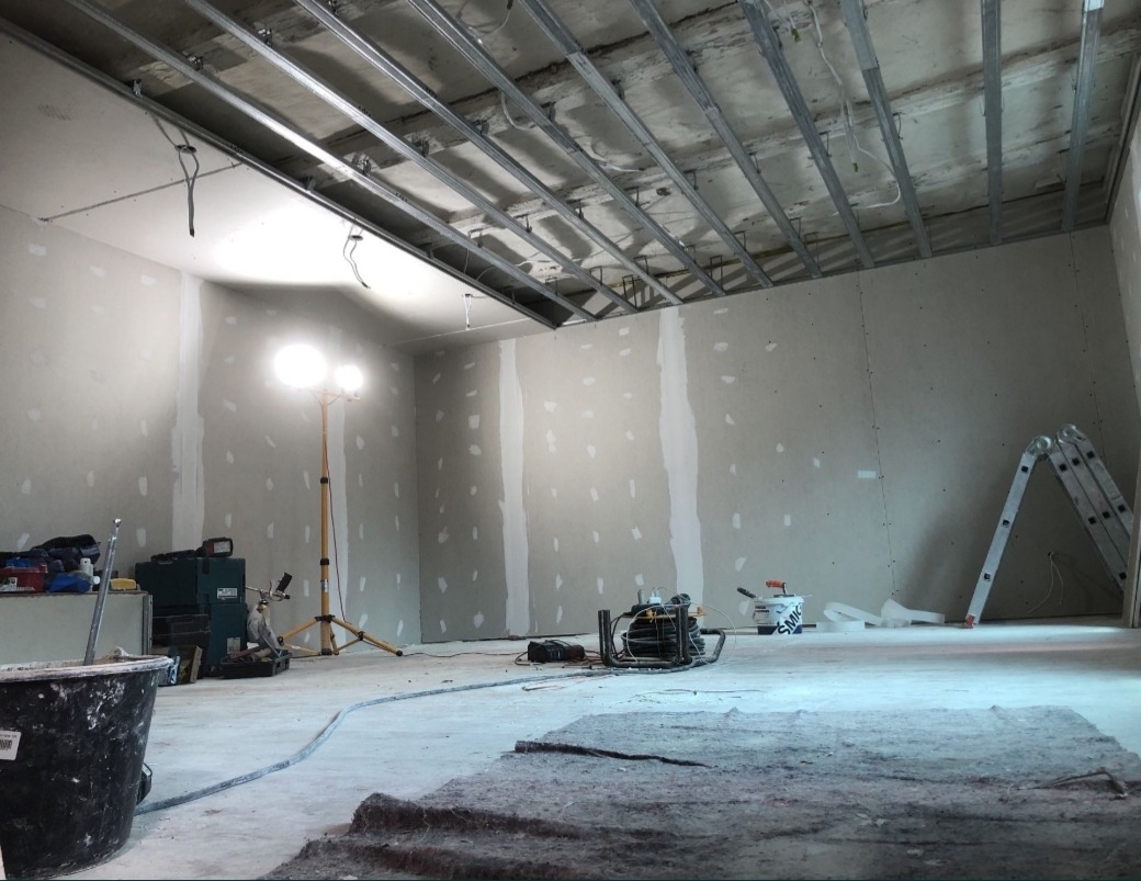 Wnętrze w trakcie prac wykończeniowych: surowy sufit z metalową konstrukcją, ściany z płyt kartonowo-gipsowych ze śladami szpachlowania, oświetlenie robocze i drabina oparta o ścianę.