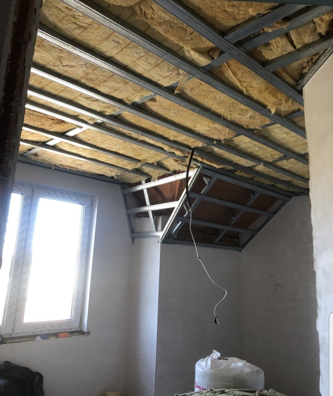 Montaż metalowej konstrukcji sufitu podwieszanego z izolacją z wełny mineralnej, widok na okno w trakcie prac wykończeniowych.