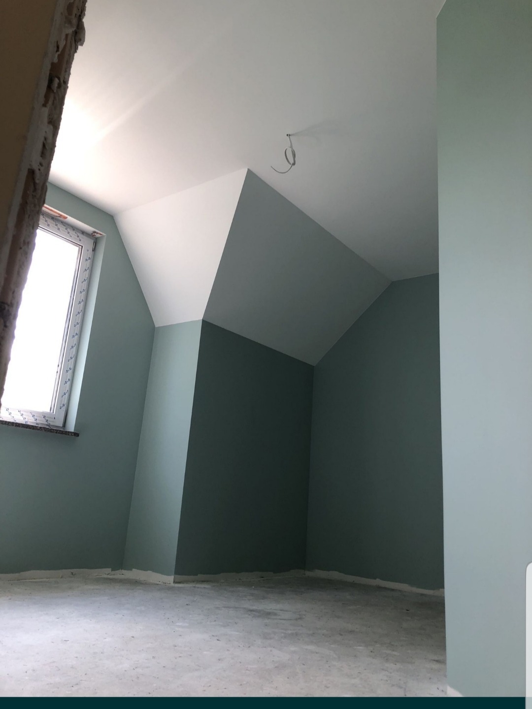 Pokój na poddaszu po malowaniu ścian na odcień zieleni, z widocznym oknem dachowym i kablem zwisającym z sufitu; surowa podłoga przed wykończeniem.