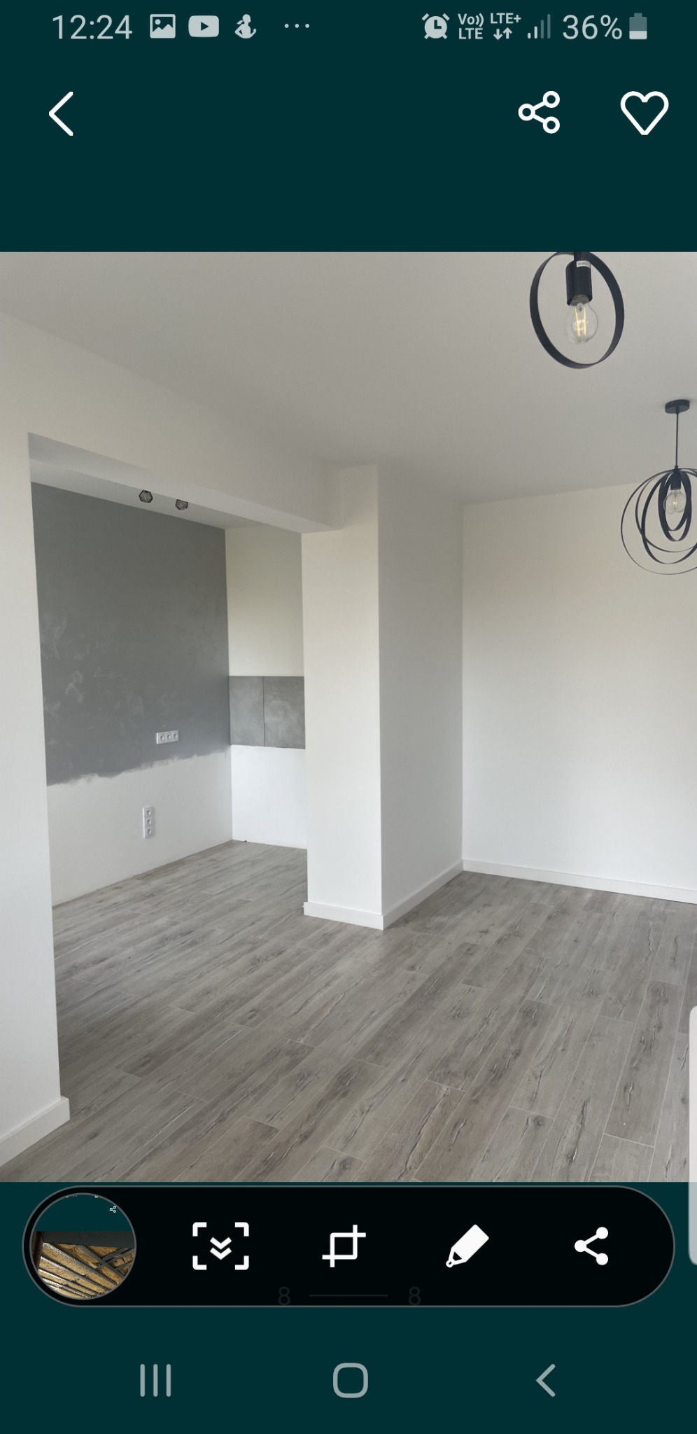 Wnętrze pokoju z szarą ścianą częściowo wykończoną, drewnopodobną podłogą, białymi ścianami i nowoczesnymi lampami wiszącymi.