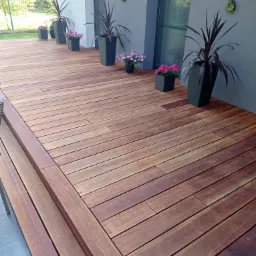 Nowoczesny taras z drewna egzotycznego, z niskimi schodami i donicami z kwiatami, oświetlony naturalnym światłem.