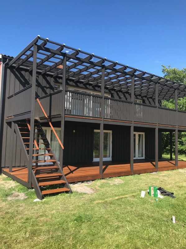 Dwukondygnacyjny budynek z ciemną elewacją, drewnianym tarasem na parterze i balkonie, zadaszony ażurową konstrukcją. Schody prowadzą na górny taras. Na trawniku widoczne narzędzia i resztki...