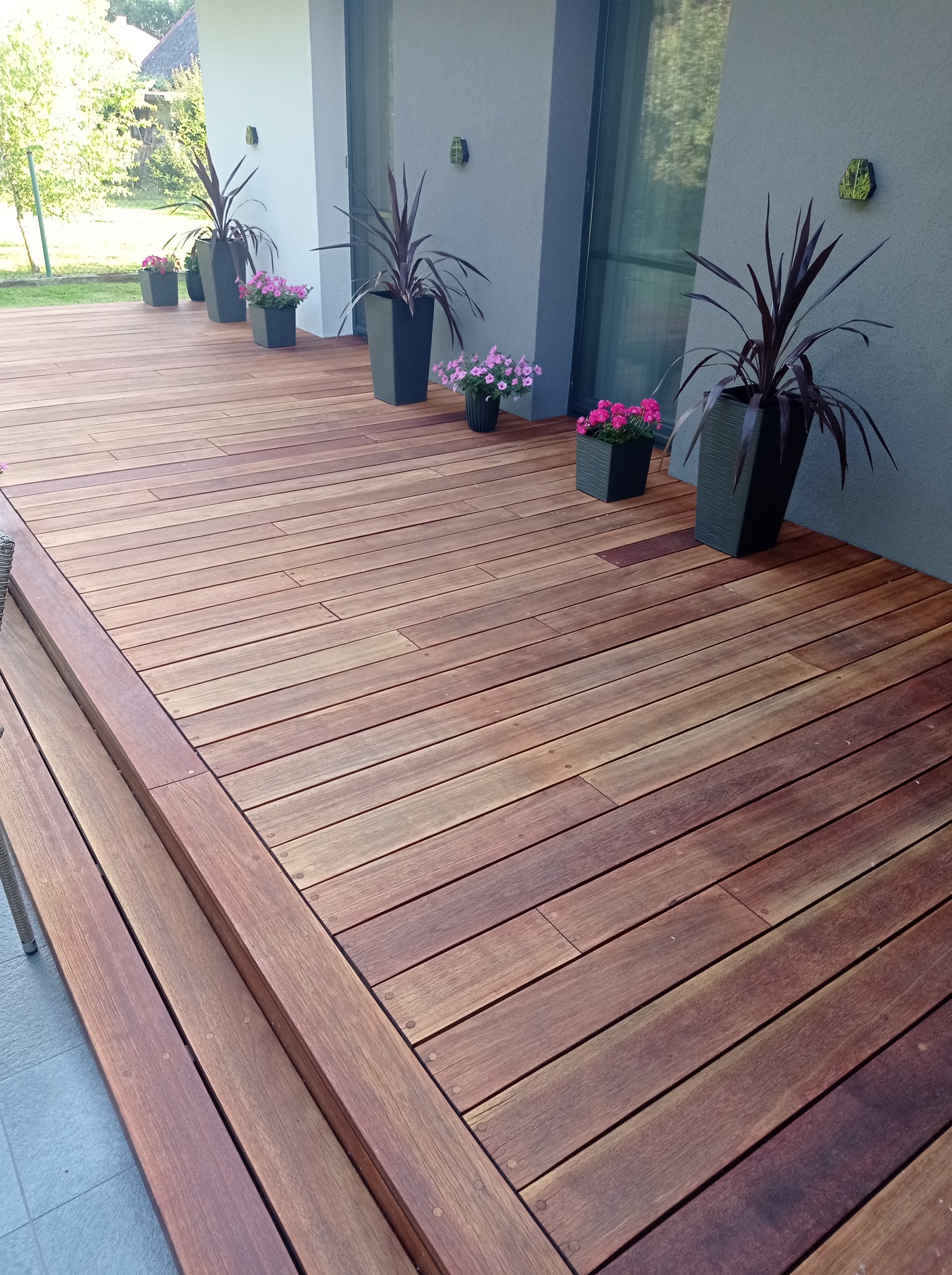 Nowoczesny taras z drewna egzotycznego, z niskimi schodami i donicami z kwiatami, oświetlony naturalnym światłem.