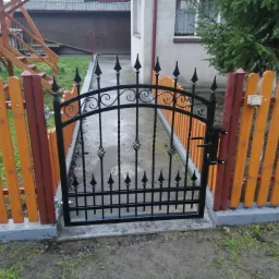 Zamykana furtka z czarnego metalu z ozdobnymi grotami, wkomponowana w pomarańczowo-brązowe drewniane ogrodzenie, na betonowej podmurówce, przed domem z zielonym iglakiem.