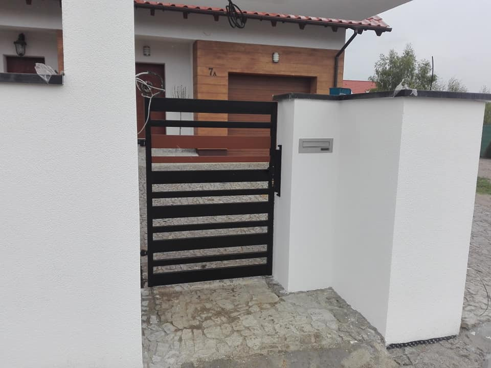 Nowoczesna, pozioma brama z czarnych, metalowych profili, zamontowana między dwoma białymi, otynkowanymi słupkami z szarym blatem, na tle domu z drewnianą elewacją.