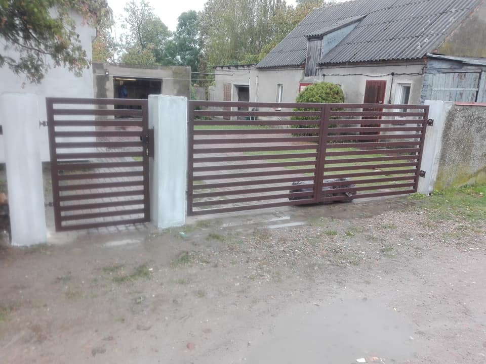 Nowoczesna brama posesyjna z poziomych profili w kolorze ciemnego brązu, osadzona na betonowych słupkach, na tle szarego, podmokłego terenu i zabudowań gospodarczych.