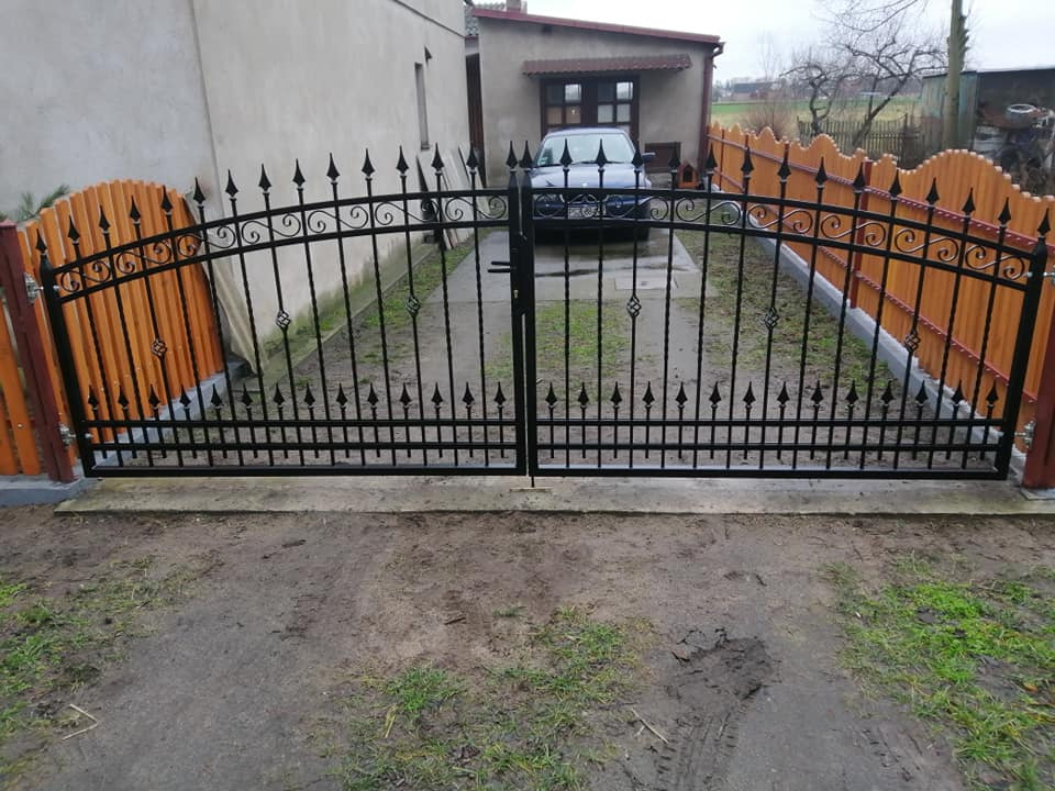 Czarna, metalowa brama wjazdowa z dekoracyjnymi elementami, osadzona w drewnianym ogrodzeniu z pionowych desek, widok na podjazd i dom w tle.