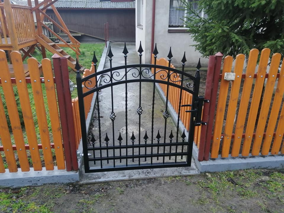 Zamykana furtka z czarnego metalu z ozdobnymi grotami, wkomponowana w pomarańczowo-brązowe drewniane ogrodzenie, na betonowej podmurówce, przed domem z zielonym iglakiem.