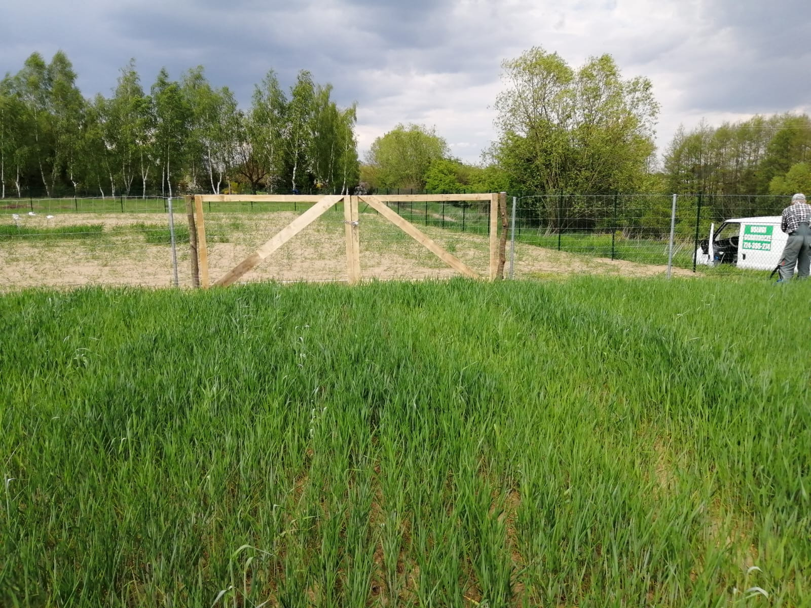Surowa drewniana brama wjazdowa z ukośnymi wzmocnieniami, zamontowana w metalowej siatce ogrodzeniowej, widok z pola zielonej trawy, częściowo zasłaniająca zaparkowany biały samochód dostawczy.