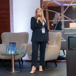 Kobieta w granatowym garniturze, z identyfikatorem na szyi, przemawia do mikrofonu na tle lustrzanej ściany z laptopem na mównicy, obok stolika z wodą i krzesłami.