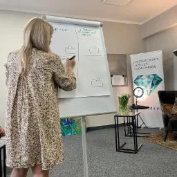 Kobieta w kwiecistej sukience rysuje markerem na flipcharcie z napisem 'OK = OK' i '2. DRUGA OSOBA TEŻ NIE OK', obok widoczny baner 'Mistrzowska Szkoła Coachingu' i wazon z białymi tulipanami.