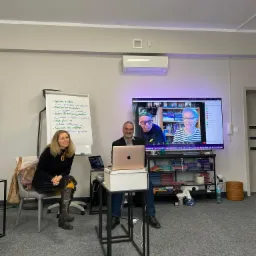 Prelegent i uczestniczka szkolenia podczas wideokonferencji, flipchart z notatkami widoczny w tle.