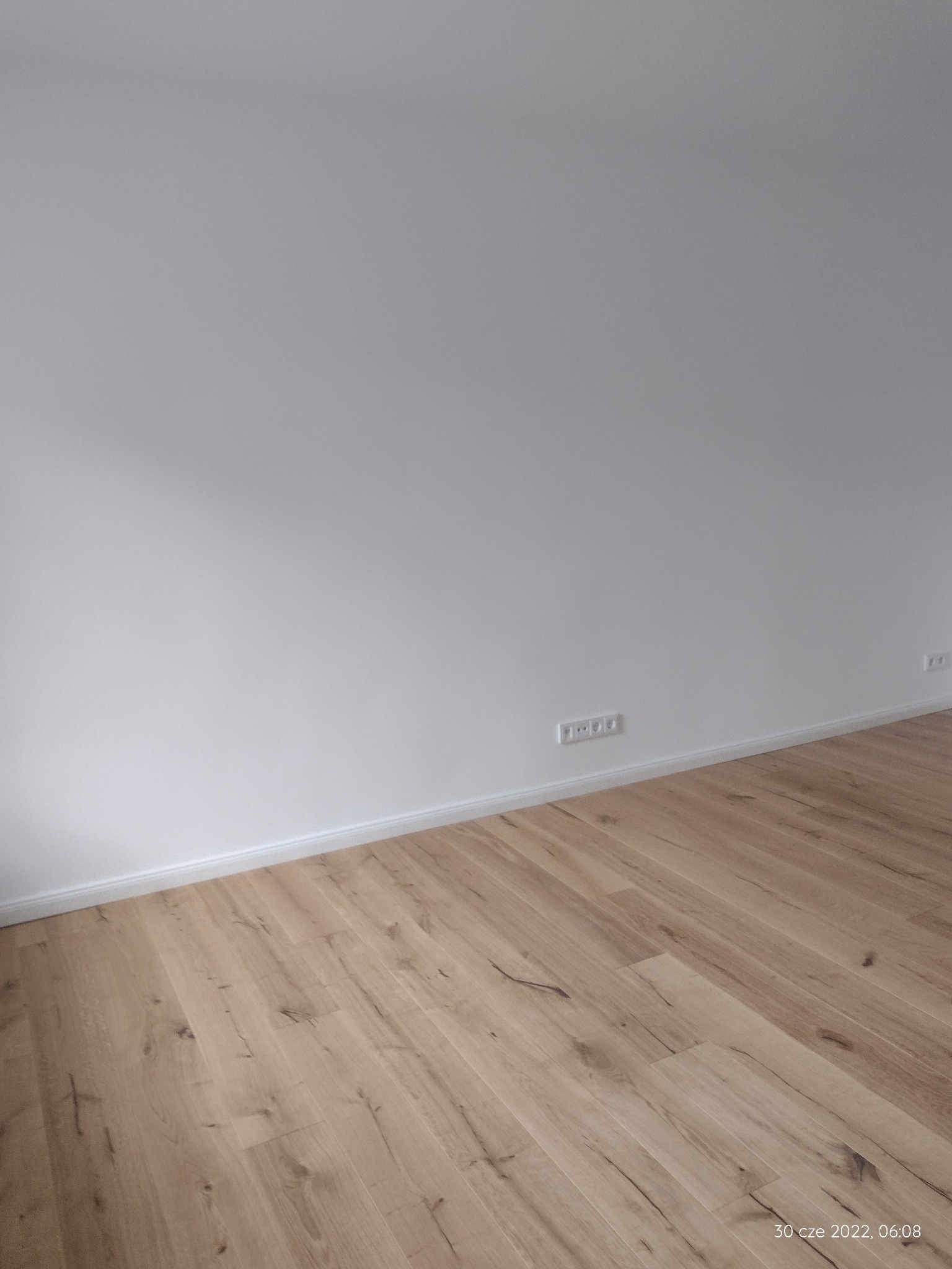 Jasna, drewniana podłoga ułożona w nowym mieszkaniu, widoczne białe ściany i listwy przypodłogowe oraz gniazdka elektryczne.