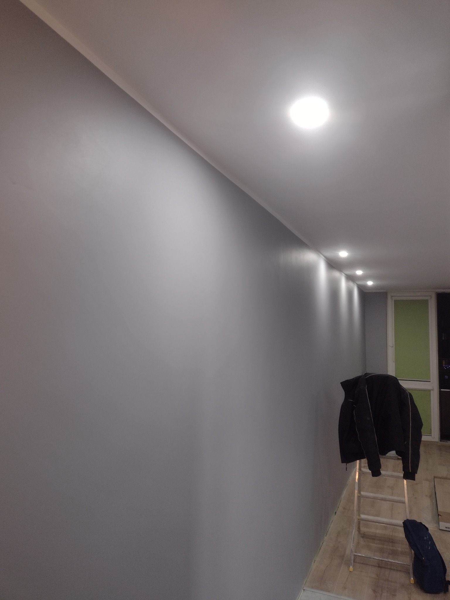 Świeżo pomalowana szara ściana z zamontowanymi lampami sufitowymi LED, drabina z kurtką w rogu pokoju, widok na okno.