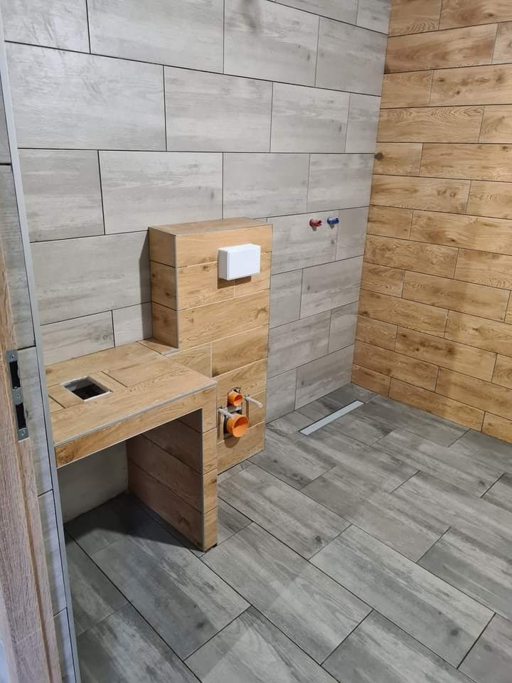 Wykończona łazienka z drewnopodobnymi płytkami w odcieniach szarości i brązu, zabudowa stelaża WC z drewnianą obudową i otworem na deskę, widoczna instalacja wodno-kanalizacyjna z rurami PCV.