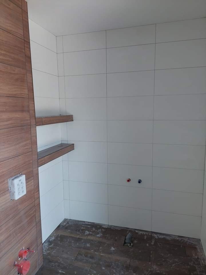 Wnętrze łazienki w trakcie prac wykończeniowych: białe płytki na ścianach, drewnopodobne półki wnękowe, wystające rury instalacji hydraulicznej, częściowo ułożona podłoga z płytek imitujących drewno.