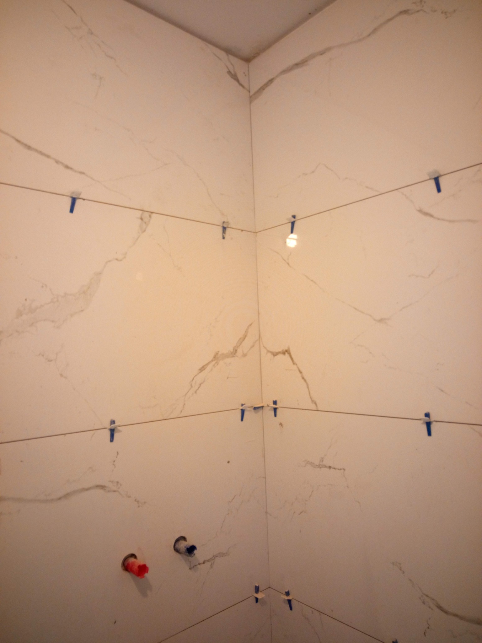 Ściana łazienki w trakcie układania płytek imitujących marmur, widoczne sznurki poziomujące, plastikowe krzyżyki dystansowe i otwory na instalacje hydrauliczne.
