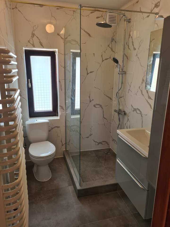 Nowoczesna łazienka z kabiną prysznicową typu walk-in, marmurowe płytki na ścianach, szara podłoga, toaleta z czarną ramą okienną i grzejnik drabinkowy.