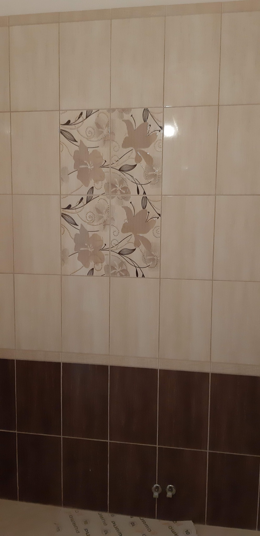 Ściana w łazience wyłożona beżowymi i brązowymi płytkami ceramicznymi, z dekoracyjnym wzorem kwiatowym na kilku płytkach.