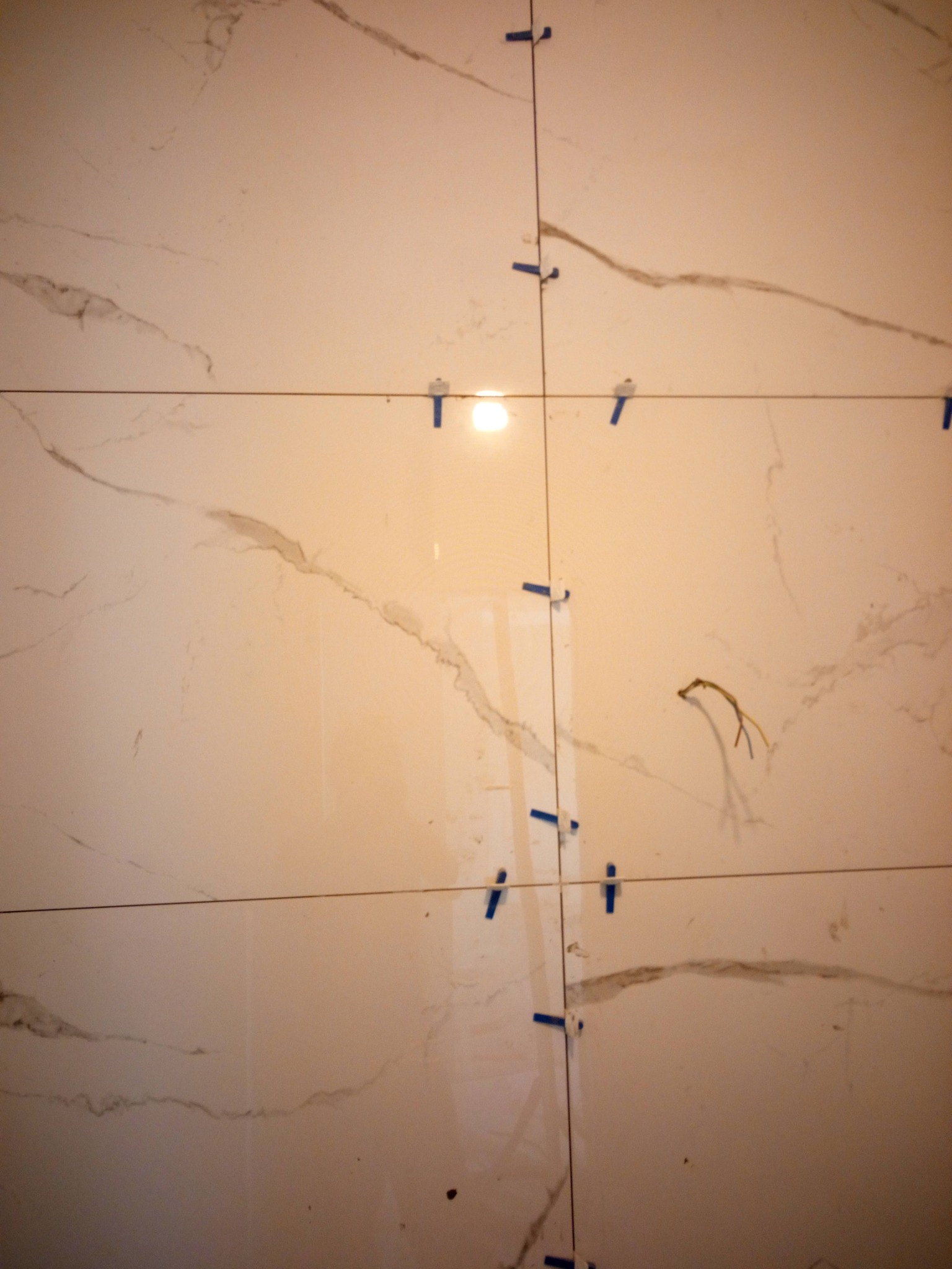 Układanie płytek imitujących marmur na ścianie, z zastosowaniem niebieskich krzyżyków dystansowych i sznurka do poziomowania, widoczne wystające przewody elektryczne.