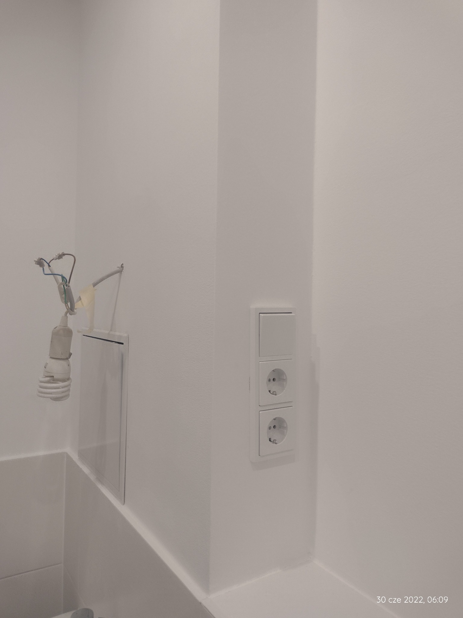 Biała łazienka w trakcie remontu, widoczne gniazdka elektryczne, włącznik światła, otwarta klapka rewizyjna i wisząca żarówka z kablami.