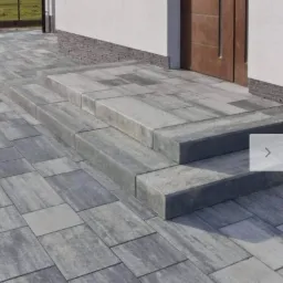 Schody wejściowe wyłożone szarą kostką brukową, prowadzące do drzwi wejściowych budynku z jasną elewacją.