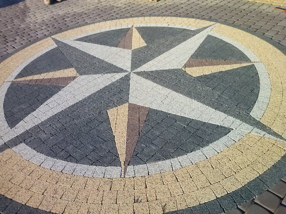 Kompas ułożony z kostki brukowej w różnych kolorach: beżowym, szarym i grafitowym, otoczony regularną kostką. Widok z góry.