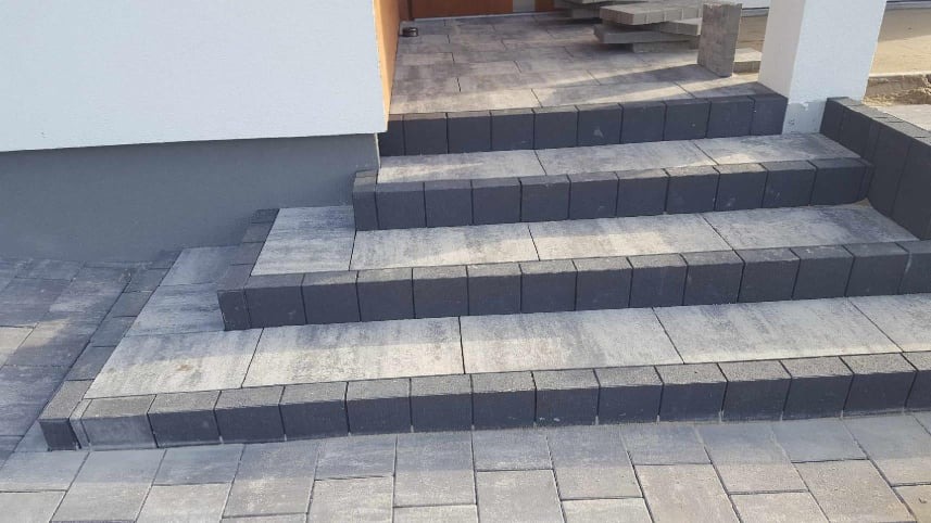 Schody wejściowe do domu wyłożone szarą kostką brukową z ciemnoszarym obramowaniem, widoczne pozostałości kostki na podjeździe