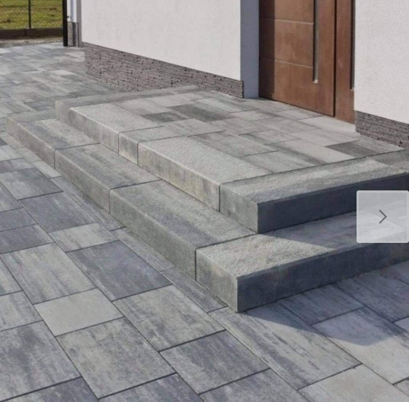 Schody wejściowe wyłożone szarą kostką brukową, prowadzące do drzwi wejściowych budynku z jasną elewacją.