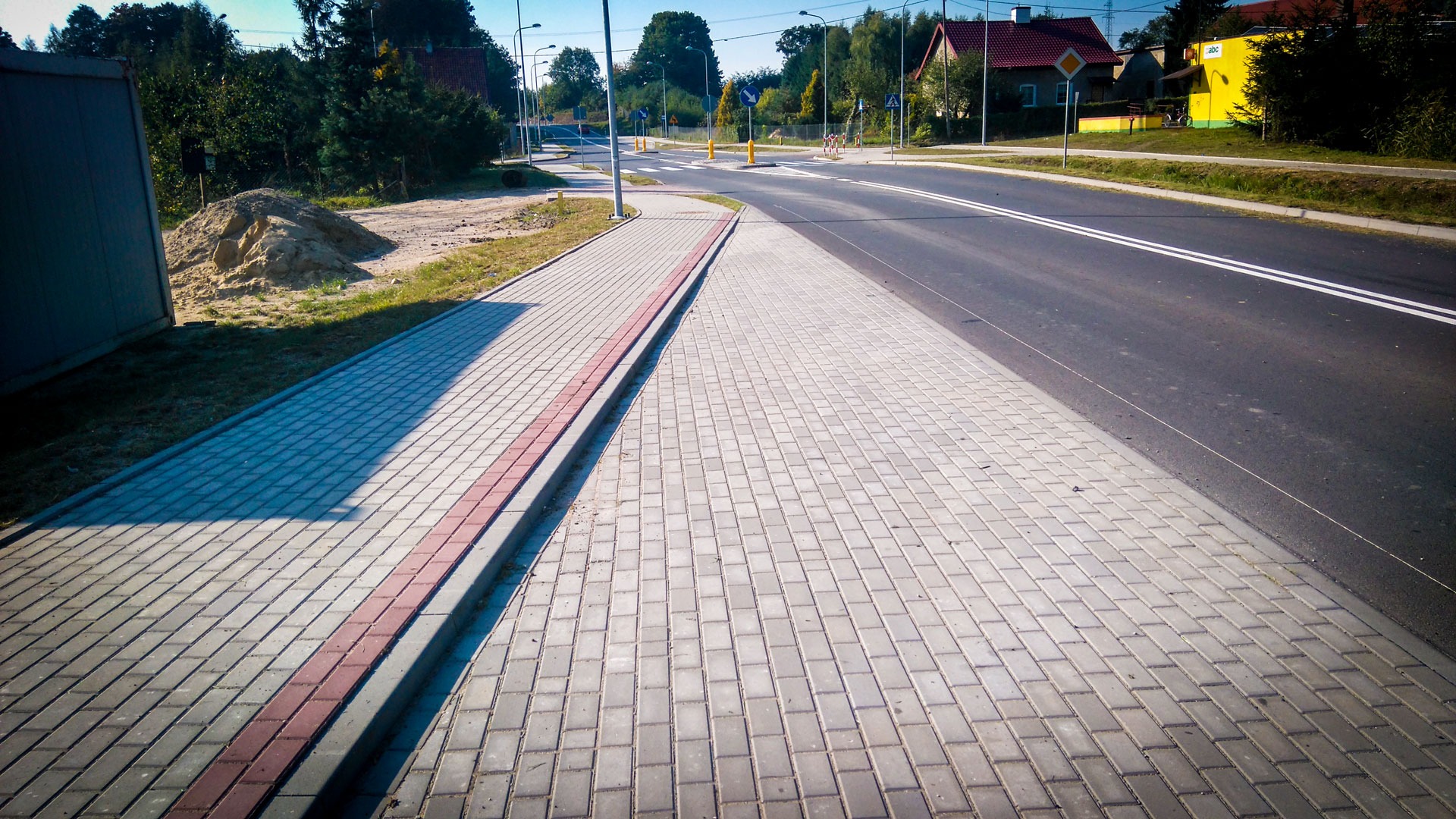 Ułożona szara kostka brukowa chodnika z czerwonym pasem oddzielającym od jezdni, widok na skrzyżowanie i zabudowania w tle.