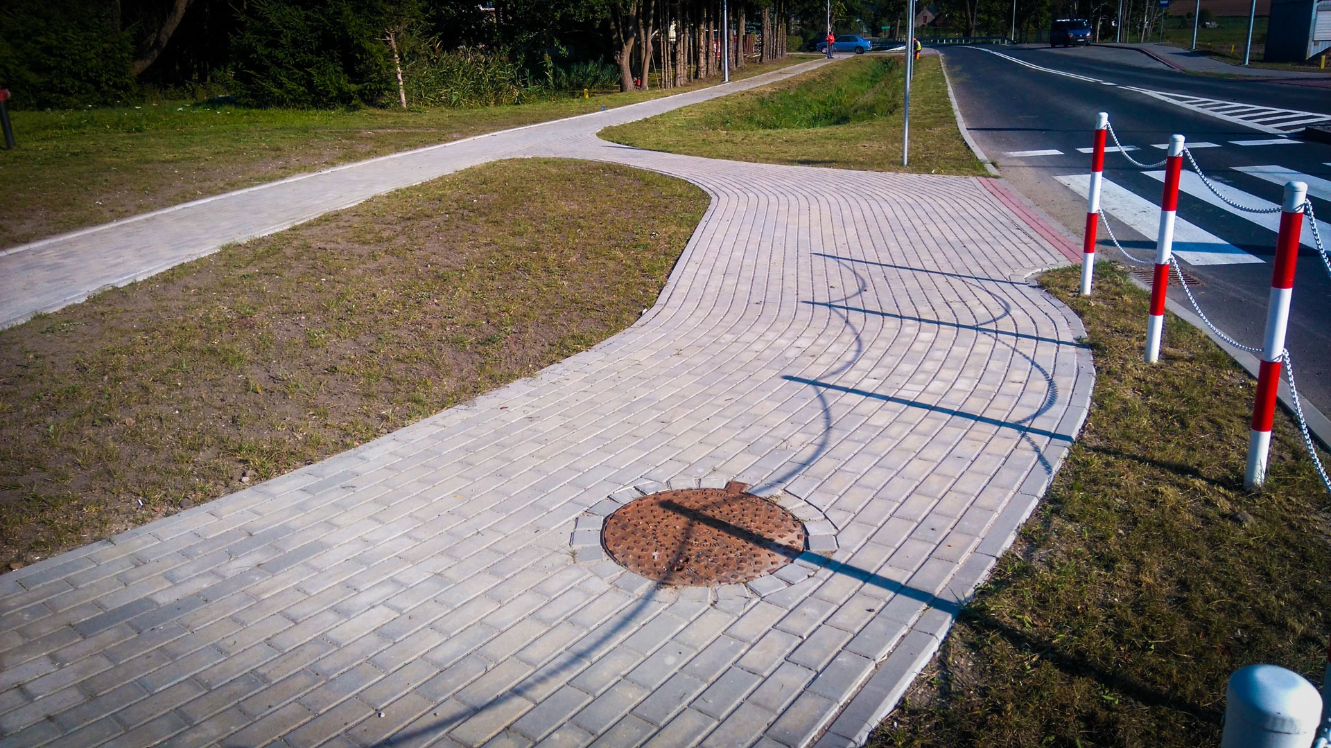 Zakrzywiona ścieżka z kostki brukowej prowadząca do przejścia dla pieszych, z widoczną studzienką kanalizacyjną i słupkami zabezpieczającymi.