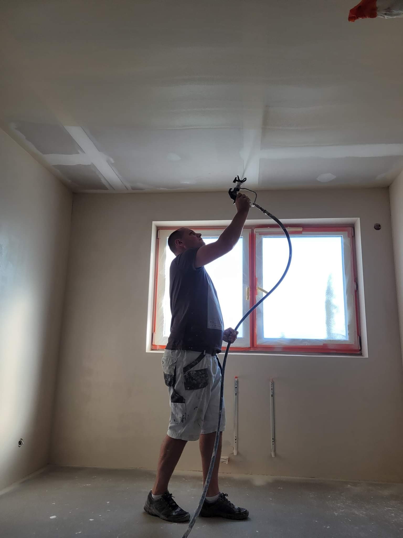 Mężczyzna w szortach maluje natryskowo sufit w pokoju z oknem zabezpieczonym folią, widoczne ślady szpachlowania na suficie.
