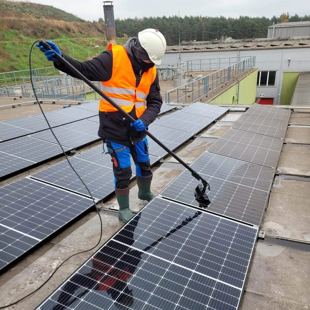 Pracownik w kasku i kamizelce odblaskowej czyści panele słoneczne za pomocą szczotki na długim wysięgniku, w tle budynek przemysłowy i drzewa.