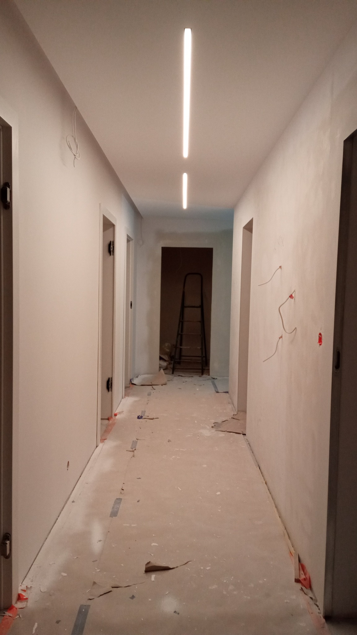 Korytarz w trakcie remontu: świeżo pomalowane ściany w odcieniach bieli, widoczne ślady po remoncie na podłodze, wystające kable ze ściany i drabina w głębi korytarza.