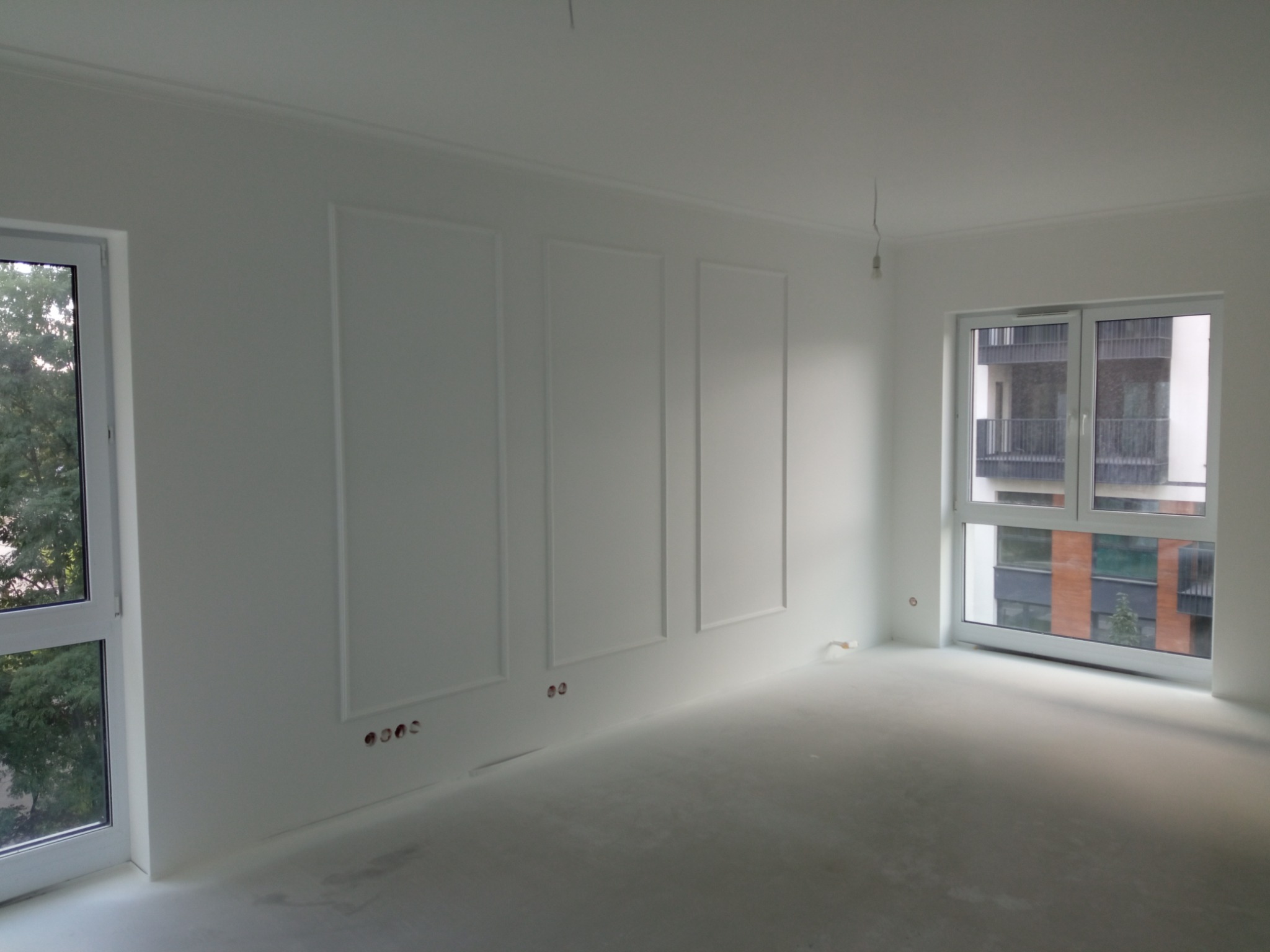 Pusty, biały pokój z trzema prostokątnymi panelami dekoracyjnymi na ścianie, widoczne gniazdka elektryczne, okna z widokiem na zieleń i sąsiedni budynek.