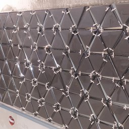 GranPlus Dmytro Libeha - Montaż błyszczących, ciemnych trójkątnych płytek na ścianie z użyciem krzyżyków dystansowych i pomarańczowych klinów poziomujących na krawędzi.