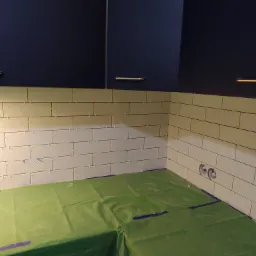 Układanie białych, podłużnych płytek z delikatnym wzorem na ścianie, widoczne krzyżyki dystansowe, zabezpieczona podłoga folią, fragment szafek wiszących.