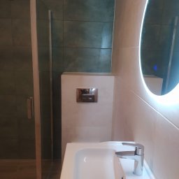 GranPlus Dmytro Libeha - Nowoczesna łazienka z kabiną prysznicową, umywalką z chromowaną baterią i okrągłym lustrem z podświetleniem LED, wykończona płytkami w odcieniach beżu i zieleni.