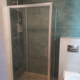 GranPlus Dmytro Libeha - Kabina prysznicowa z przesuwnymi drzwiami, chromowana armatura, zielone płytki na ścianach i drewnopodobna podłoga w łazience.