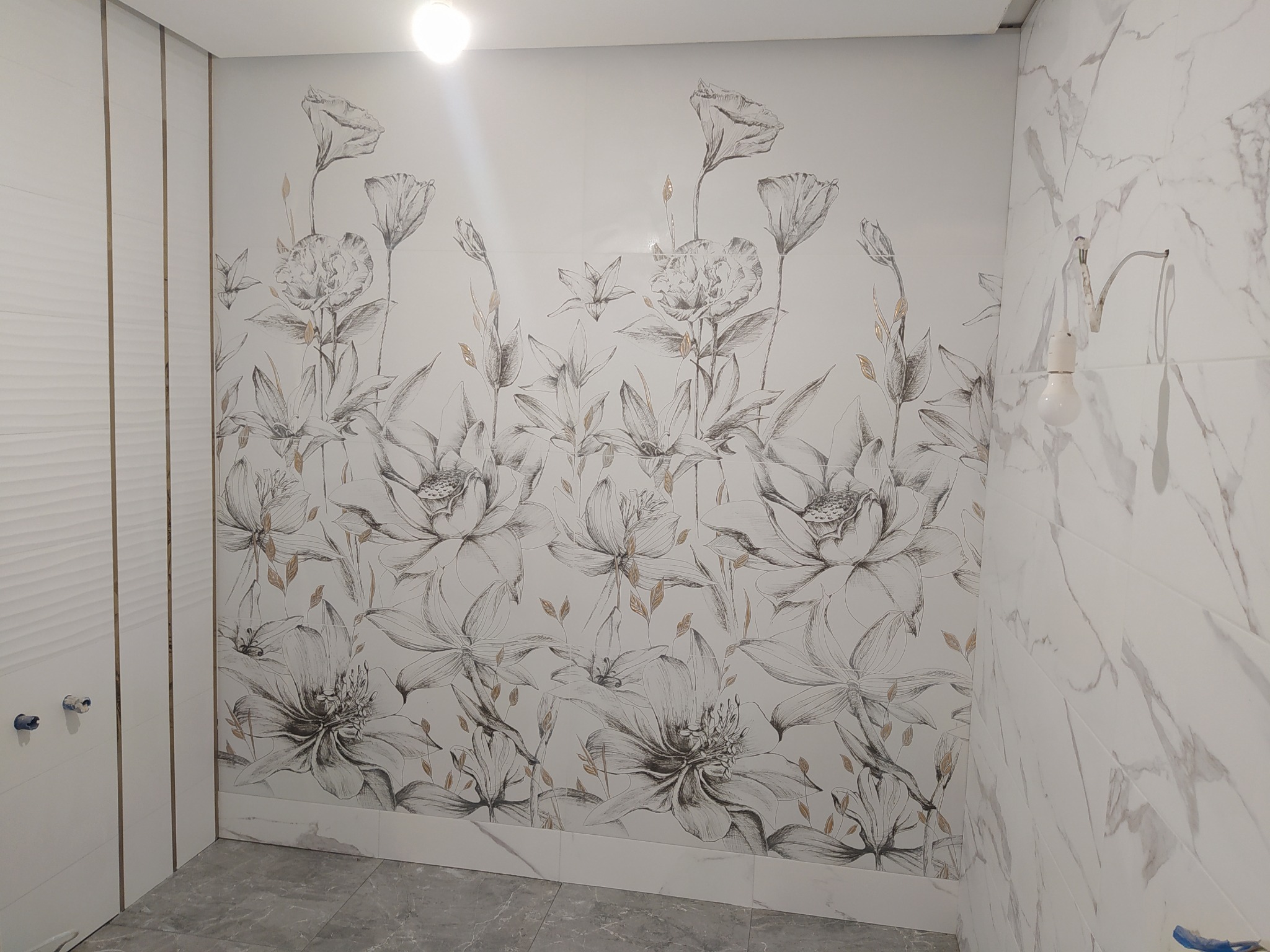 Ściana w łazience wyłożona białymi płytkami z motywem kwiatowym, sąsiadująca z białą ścianą z pionowymi żłobieniami i wystającymi elementami instalacji wodnej, oświetlona żarówką.