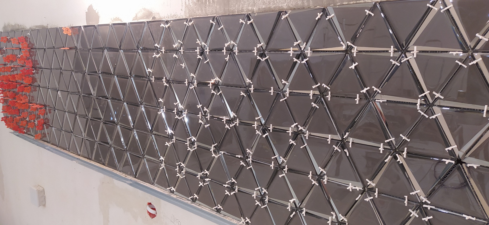 Montaż błyszczących, ciemnych trójkątnych płytek na ścianie z użyciem krzyżyków dystansowych i pomarańczowych klinów poziomujących na krawędzi.