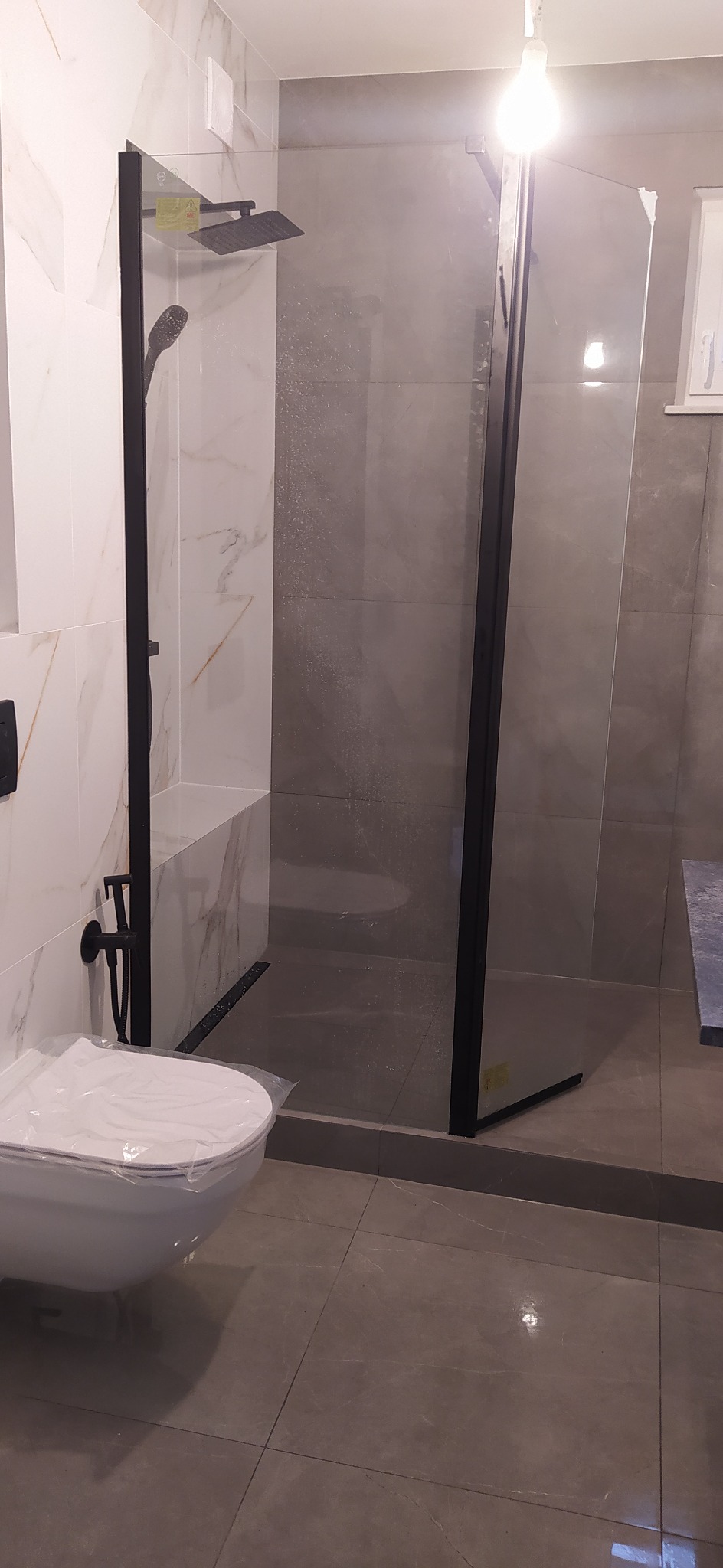 Nowoczesna łazienka z kabiną prysznicową typu walk-in z czarnymi ramami, deszczownicą i baterią podtynkową w kolorze czarnym, oraz toaletą z przykryciem z folii ochronnej.