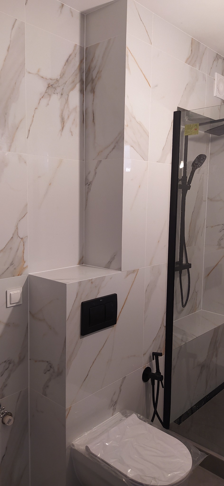 Wykończona łazienka z białymi płytkami imitującymi marmur, czarna armatura, podwieszana toaleta z przyciskiem spłukującym Roca, kabina prysznicowa z czarną ramą.