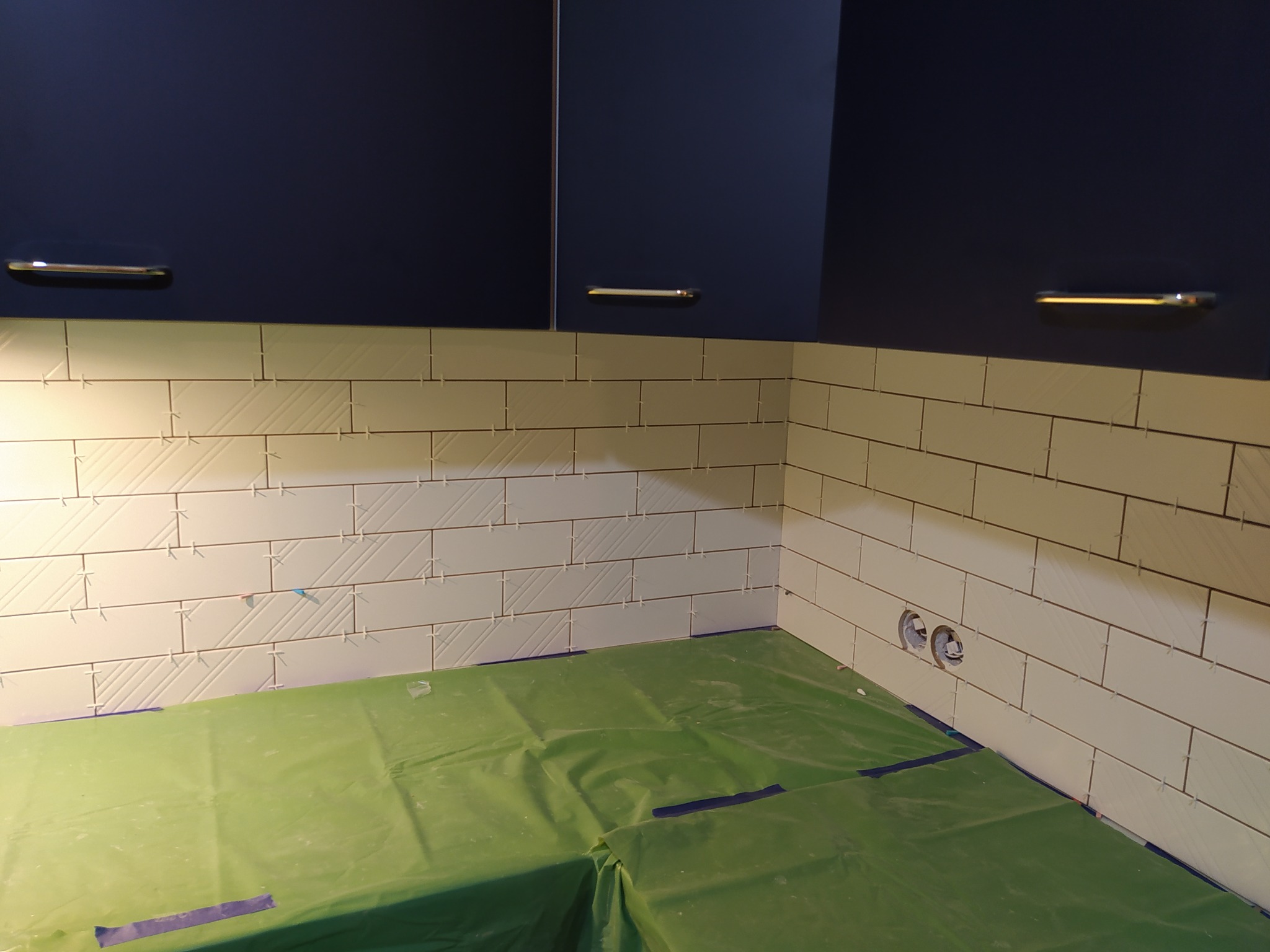 Układanie białych, podłużnych płytek z delikatnym wzorem na ścianie, widoczne krzyżyki dystansowe, zabezpieczona podłoga folią, fragment szafek wiszących.