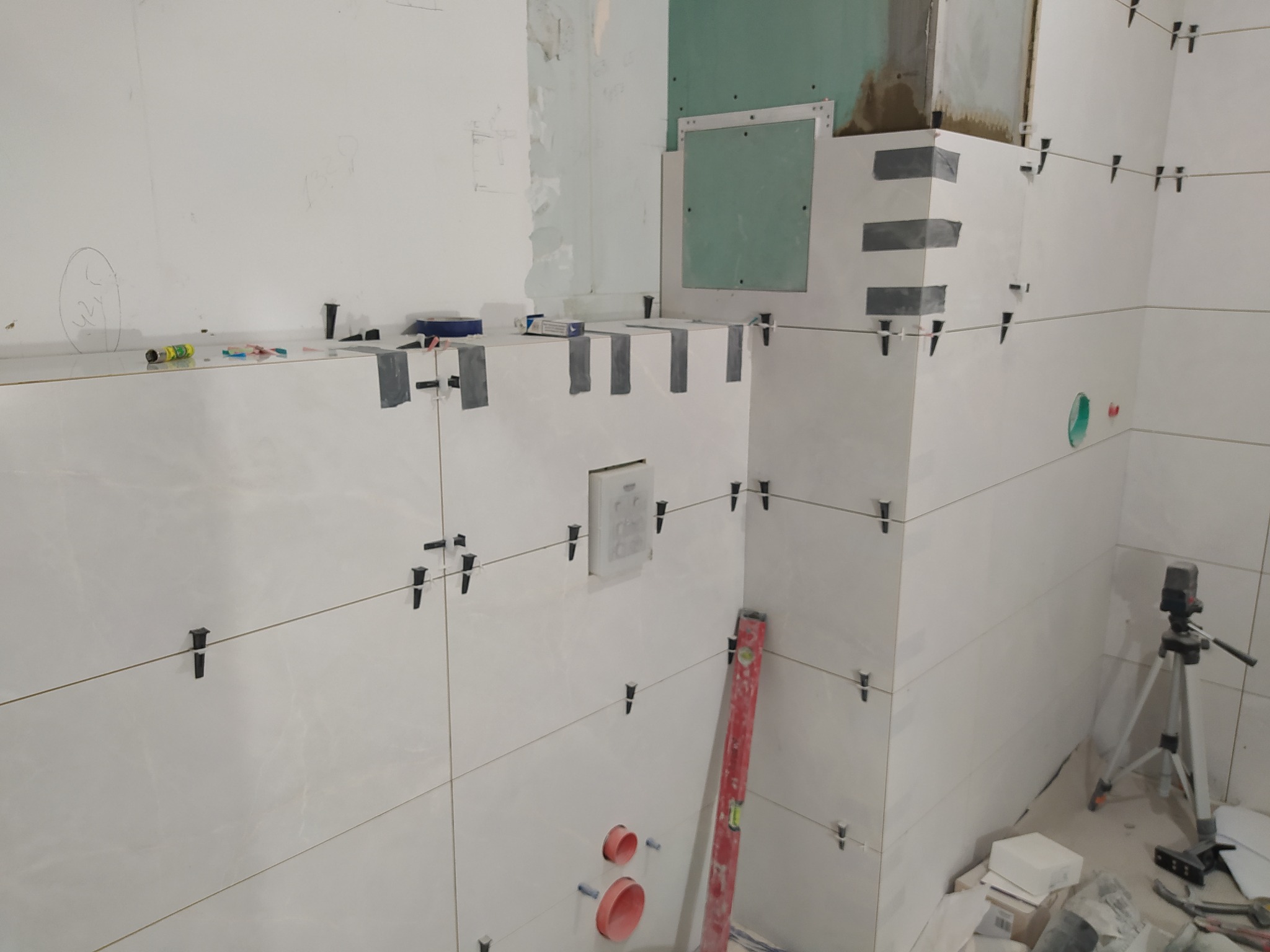 Białe płytki ceramiczne układane w łazience, widoczne krzyżyki dystansowe i poziomica laserowa na statywie.