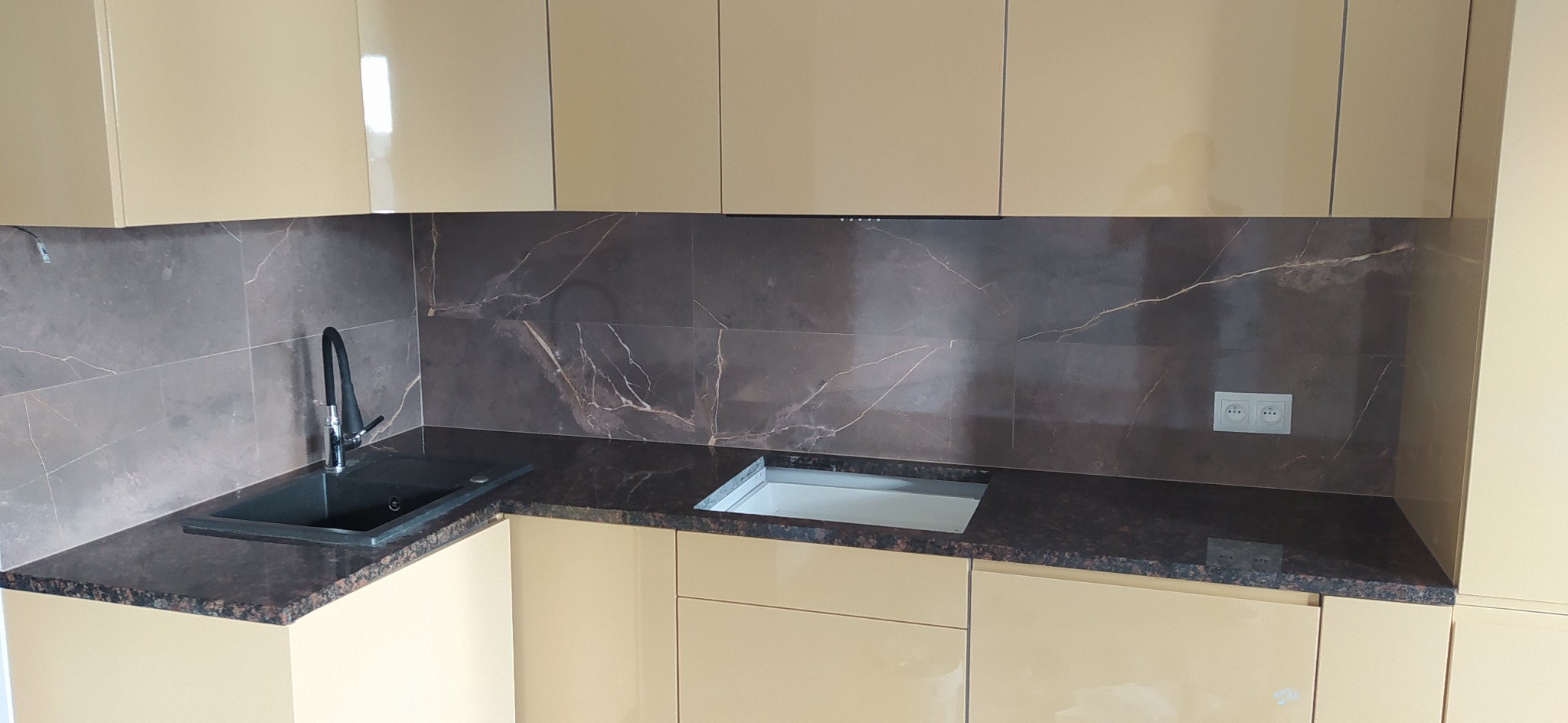 Beżowe szafki kuchenne z ciemnobrązowym blatem i marmurową ścianą nad blatem, czarny zlew z zakrzywioną baterią, gniazdka elektryczne.