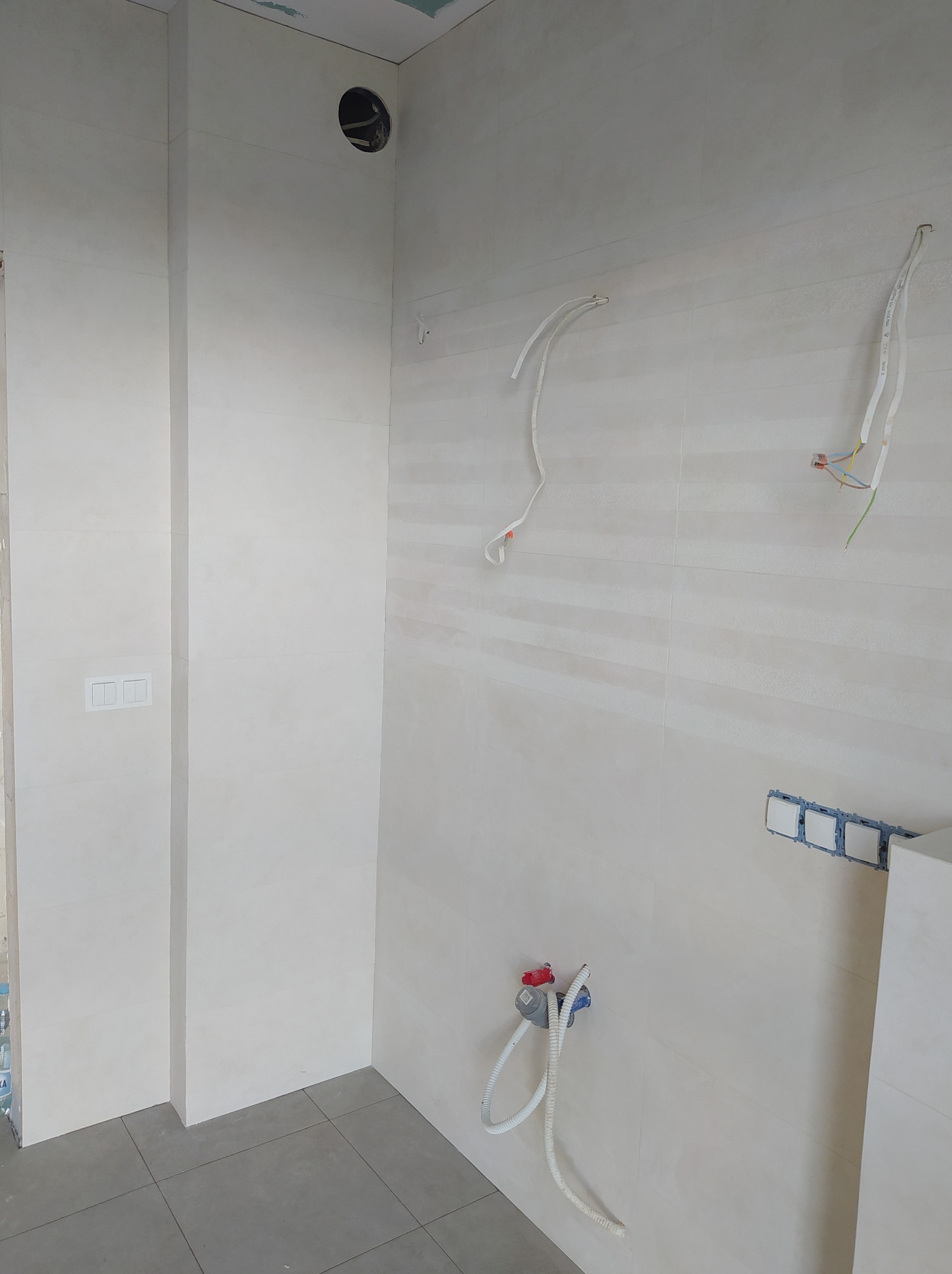 Wnętrze łazienki w trakcie prac wykończeniowych: ułożone płytki ścienne w odcieniach beżu, widoczne wystające przewody elektryczne i instalacja wodna, szara podłoga z płytek, otwór w ścianie...