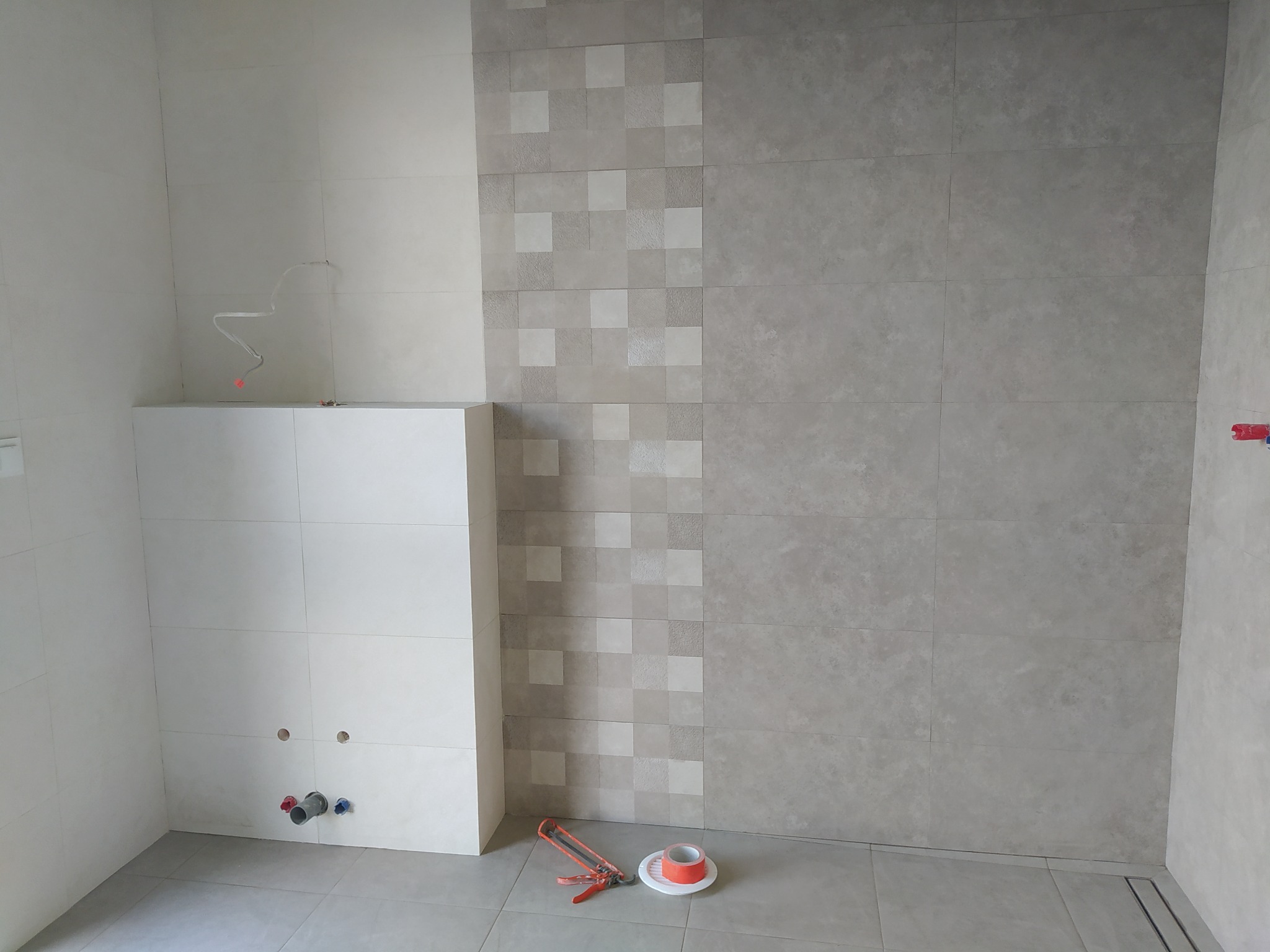 Ściana łazienki w trakcie układania płytek, widoczne przyłącza wodne, narzędzia glazurnicze i fragment dekoracyjnej mozaiki.