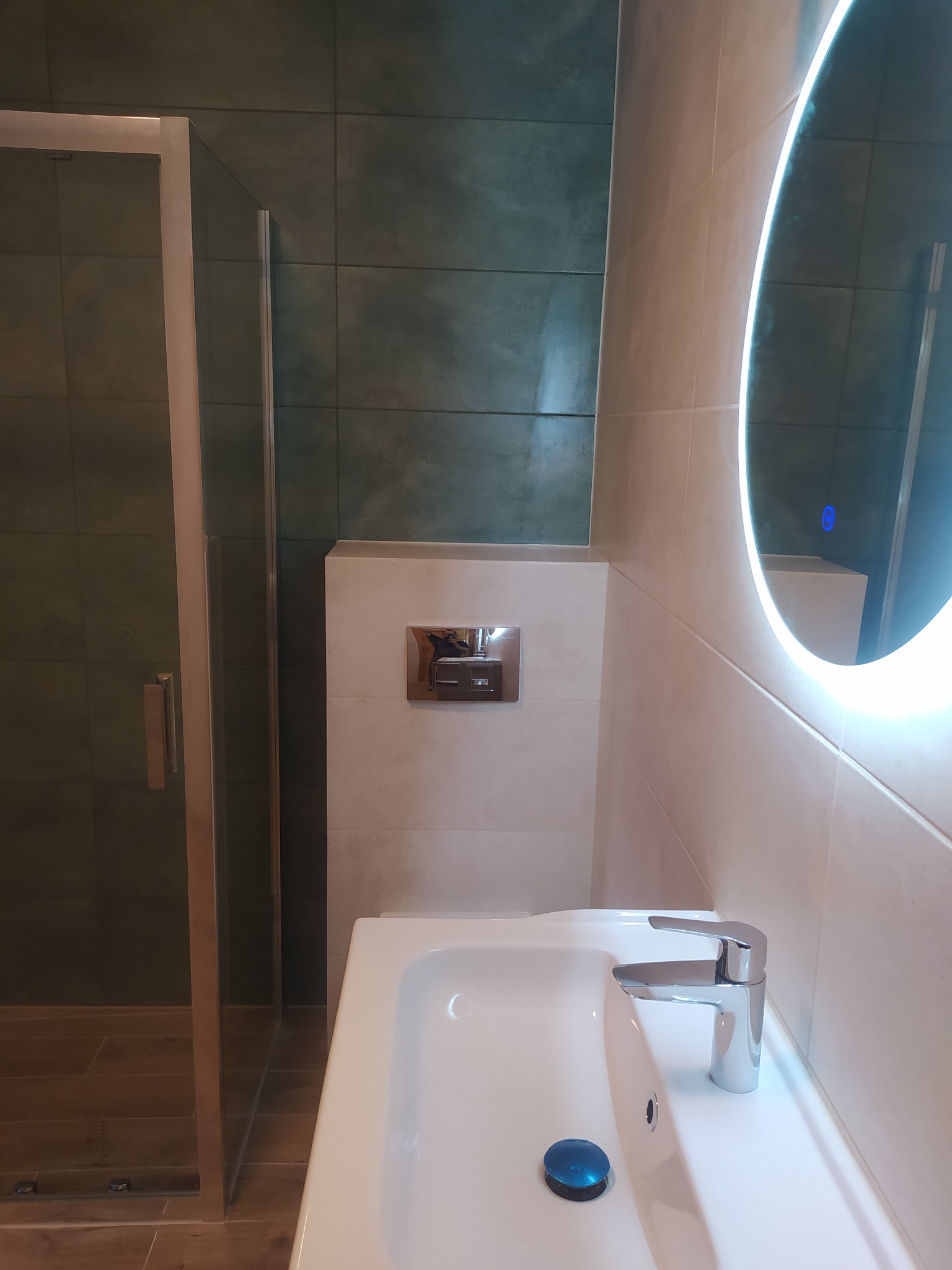 Nowoczesna łazienka z kabiną prysznicową, umywalką z chromowaną baterią i okrągłym lustrem z podświetleniem LED, wykończona płytkami w odcieniach beżu i zieleni.