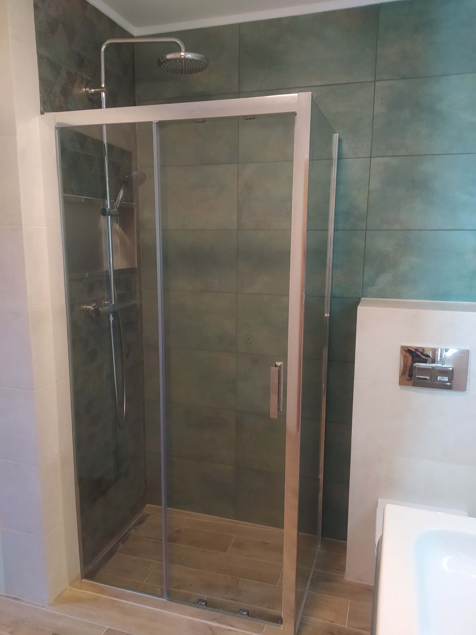 Kabina prysznicowa z przesuwnymi drzwiami, chromowana armatura, zielone płytki na ścianach i drewnopodobna podłoga w łazience.